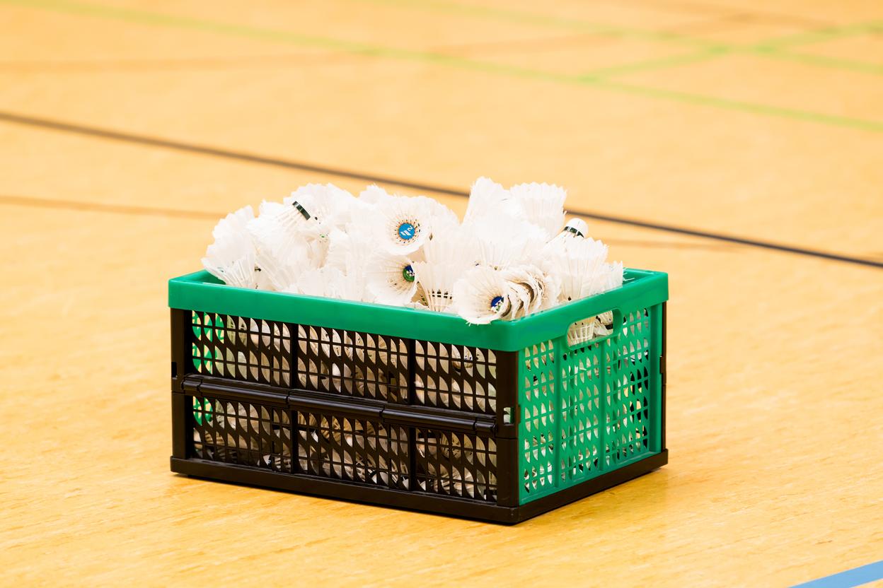 Ein grüner Korb mit vielen weißen Federbällen steht auf einem Holzfußboden in einer Sporthalle.
