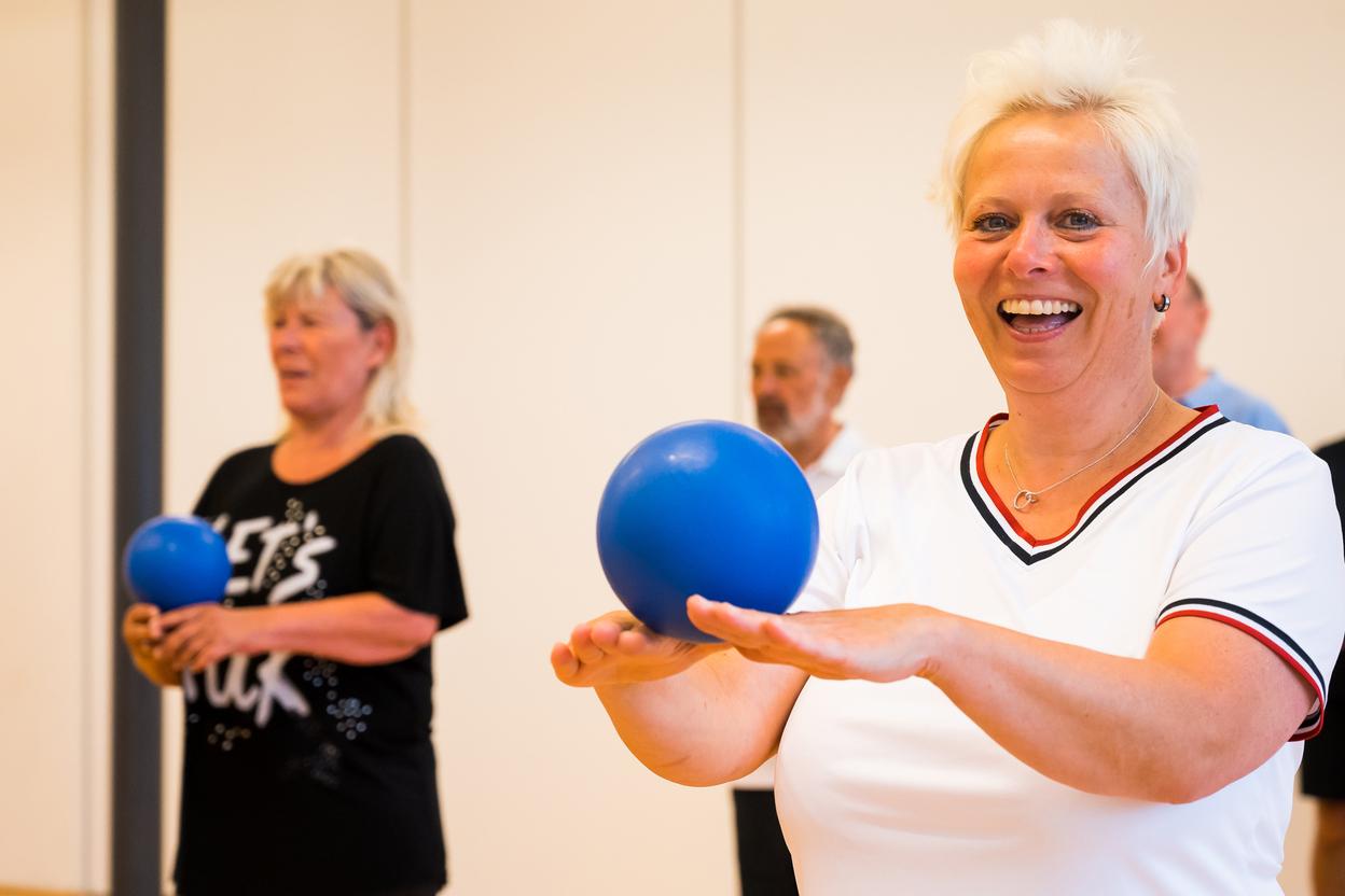 Gruppe älterer Menschen bei einer Übung mit blauen Bällen in einem hellen Raum. Fokus auf lächelnde Frau vorne.