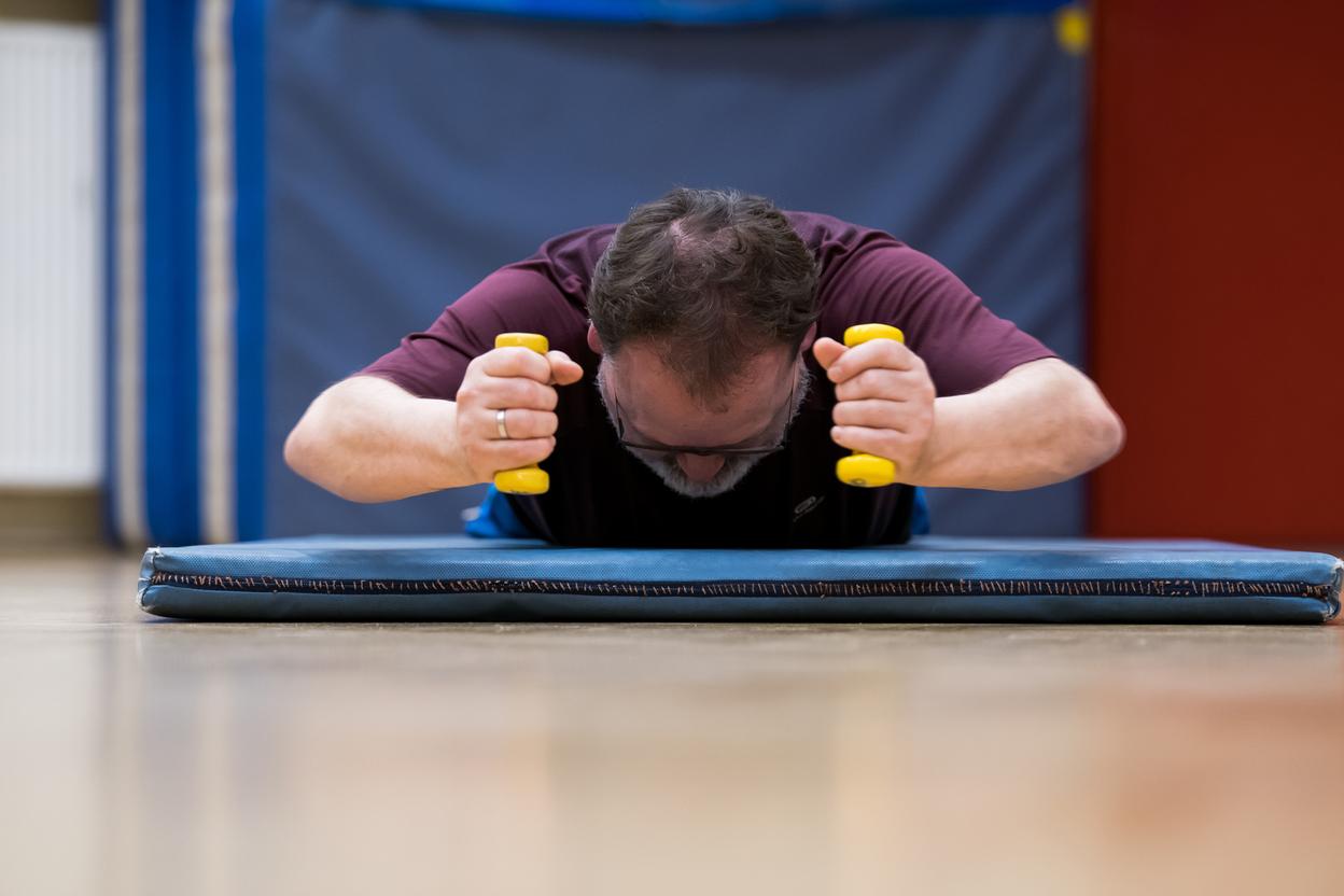 Mann trainiert auf einer Matte mit gelben Hanteln, Kopf nach unten, in einem Sportraum mit blauen Wänden.