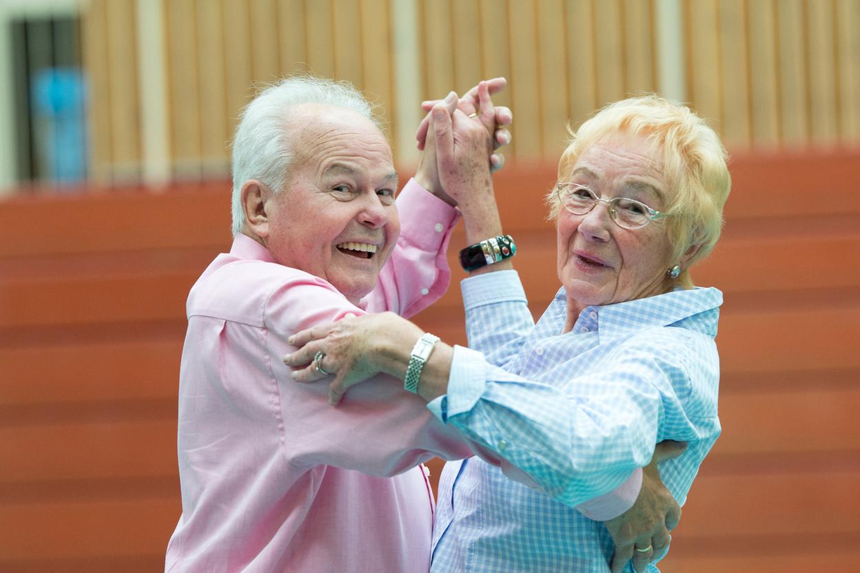 Ein älteres Paar tanzt lächelnd in einer Turnhalle, Kleidung in sanften Farben. Holzpaneele im Hintergrund.