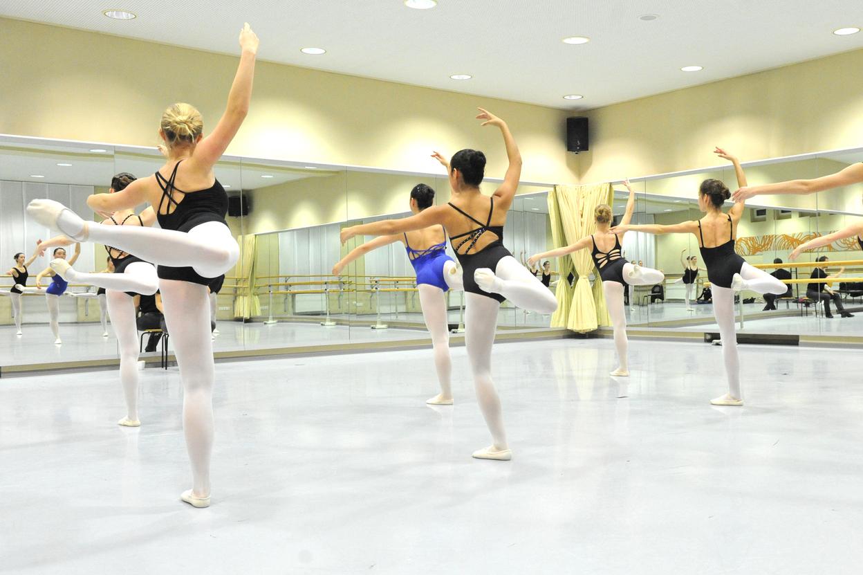 Gruppe von Balletttänzerinnen in Schwarz und Blau übt in einem Spiegelraum mit Holzballettstangen.