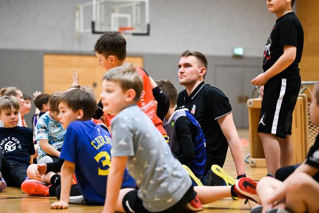 Gruppe von Kindern sitzt auf dem Boden einer Sporthalle, während ein Trainer aufmerksam auf sie schaut.